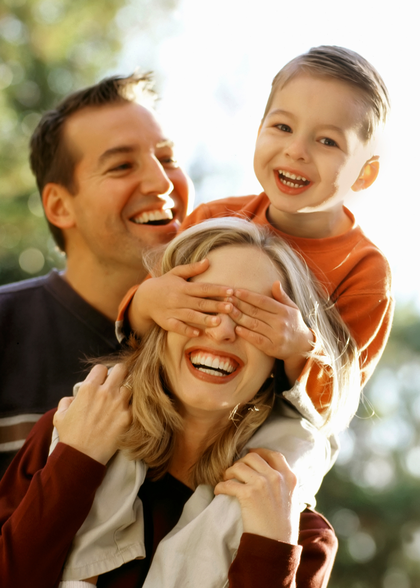 Happy local family. Son on mom's shoulders. both with big smiles. Dad looks on smiling. Outdoors.